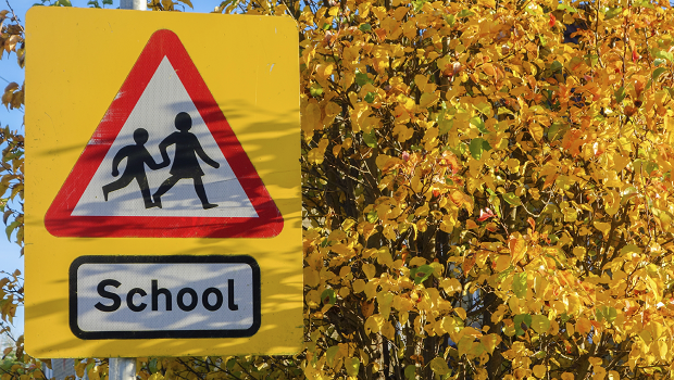 a school road sign in the forefront, trees in the backgroudn, the leaves with a yellow tinge suggesting early onset signs of autumn