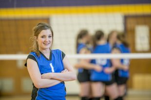A teenage girl is standing in a gym wearing a volleyball uniform and is smiling and looking at the camera. Her teammates are behind her in a team huddle.