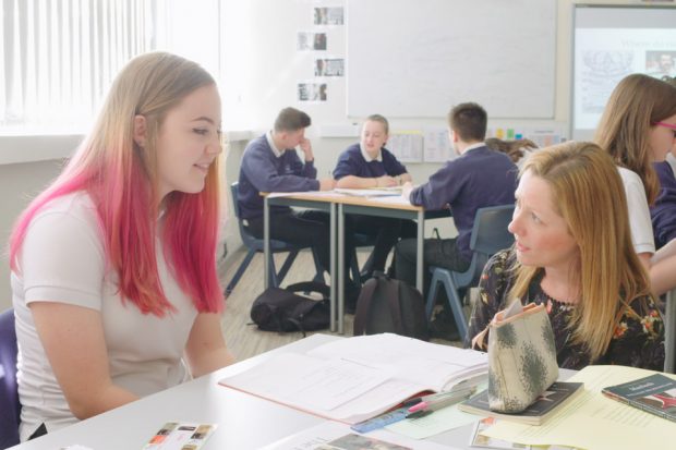 female pupil with pink hair sits next to teacher who appears to be explaining a piece of work